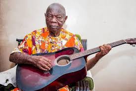Veteran highlife musician Mike Ejeagha playing guitar, wearing traditional attire and a warm, gentle expression